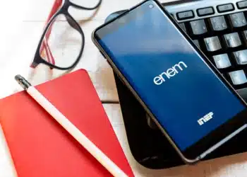 Smartphone exibindo o logo do ENEM sobre um teclado, com materiais de estudo (caderno, lápis e óculos) ao lado, simbolizando a preparação digital e o foco nos temas mais cobrados de Matemática e Física para 2025.