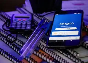 Celular exibindo o aplicativo do ENEM sobre pilhas de cadernos, acompanhado de duas canetas azuis e uma calculadora, ilustrando o foco na organização e nas ferramentas digitais e de estudo necessárias para a preparação para o ENEM 2025.
