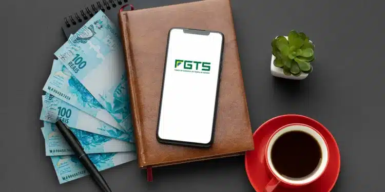 "Notas de 100 reais e celular com logo do FGTS sobre uma mesa com café, caderno e planta. Imagem relacionada ao saque-aniversário do FGTS.