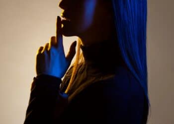 Perfil de mulher com olhos fechados, luz azul e amarela iluminando o rosto e cabelo, fazendo gesto de silêncio