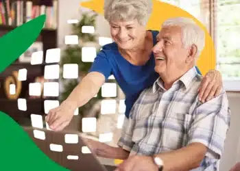 Idoso sorrindo com sua esposa, ambos usando um laptop e discutindo sobre aposentadoria, em um ambiente doméstico.