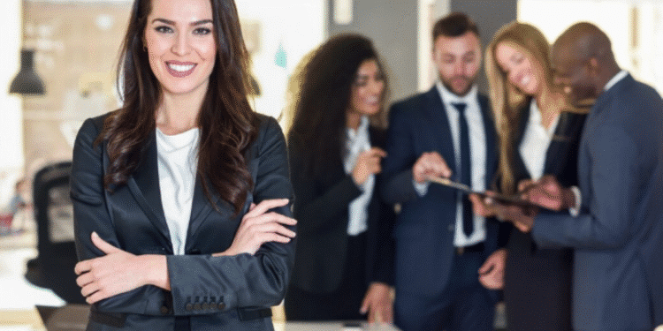 Mulher sorridente e confiante de terno preto em destaque com equipe discutindo ao fundo em ambiente corporativo