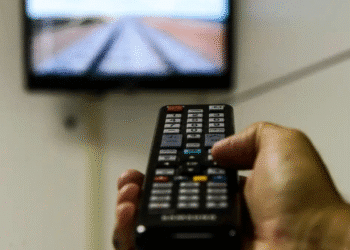 Mão segurando controle remoto apontado para televisão com imagem desfocada