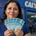Mulher segurando cédulas de R$ 100 em frente a uma agência da Caixa, representando pagamentos do Bolsa Família em julho.