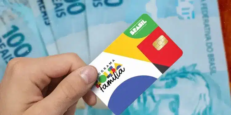 Cartão do Bolsa Família com cédulas de 100 reais ao fundo, simbolizando o programa de transferência de renda do Brasil.