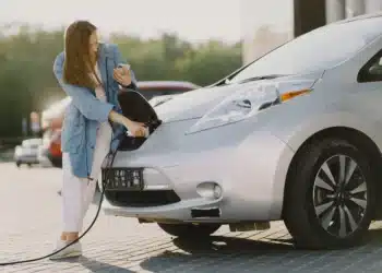Mulher carregando um carro elétrico enquanto consulta o celular, em um ambiente ao ar livre, ilustrando o cotidiano de quem possui um veículo elétrico.