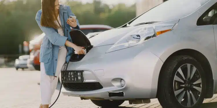 Mulher carregando um carro elétrico enquanto consulta o celular, em um ambiente ao ar livre, ilustrando o cotidiano de quem possui um veículo elétrico.