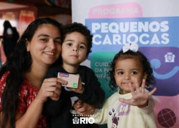 Mulher com duas crianças segurando cartão do programa Pequenos Cariocas em frente a banner oficial
