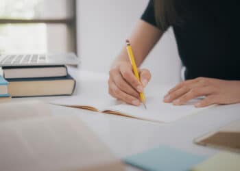 Pessoa estudando e escrevendo em um caderno com livros e notebook ao fundo.