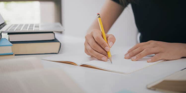 Pessoa estudando e escrevendo em um caderno com livros e notebook ao fundo.