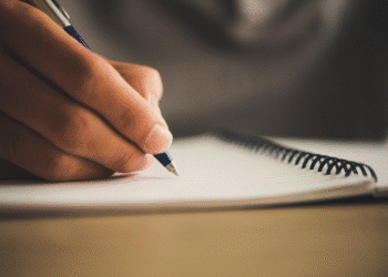 Mão segura caneta azul escrevendo em caderno espiral aberto sobre mesa de madeira