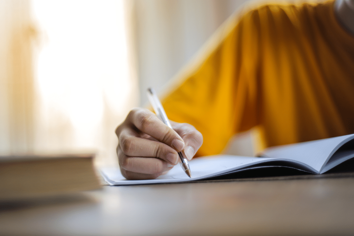 Mão segurando caneta escrevendo em caderno aberto sobre mesa com luz natural ao fundo