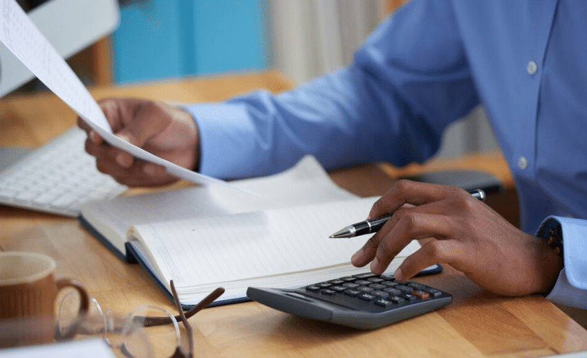 Mãos de pessoa usando caneta e calculadora sobre mesa de madeira com caderno aberto e papel na mão