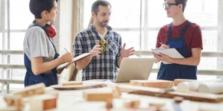 Imagem ilustrativa de profissionais em treinamento em uma oficina técnica, discutindo projetos com o uso de computadores e material de trabalho.