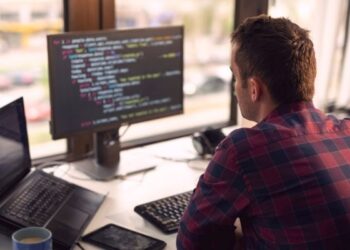 Jovem desenvolvedor de software de costas, trabalhando em frente a dois monitores exibindo código e utilizando um laptop em um escritório moderno, ilustrando o profissional e a carreira de tecnologia.