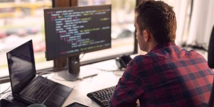 Jovem desenvolvedor de software de costas, trabalhando em frente a dois monitores exibindo código e utilizando um laptop em um escritório moderno, ilustrando o profissional e a carreira de tecnologia.