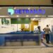 Fachada de um posto de atendimento do DETRAN RJ dentro de um shopping ou centro comercial. Pessoas aguardam ou conversam em balcões de atendimento, destacando-se a placa azul com o logo e o nome “DETRAN.RJ” em letras maiúsculas.