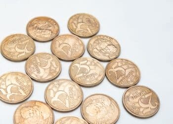 Close-up de várias moedas brasileiras de 25 centavos espalhadas, com destaque para as datas e o número. A imagem ilustra as peças que podem ser raras por erro de cunhagem ou baixa tiragem, como as edições de 2016 e 2021.