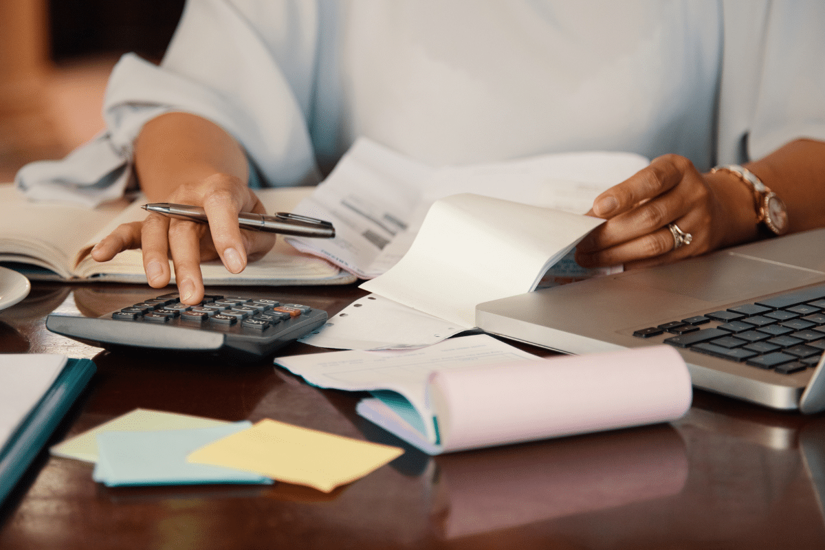 Pessoa usando calculadora e manuseando notas fiscais com notebook e papéis coloridos na mesa