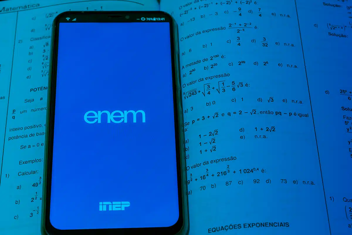 Enem 2025 Celular com tela do aplicativo ENEM aberto sobre livro de exercícios de matemática.