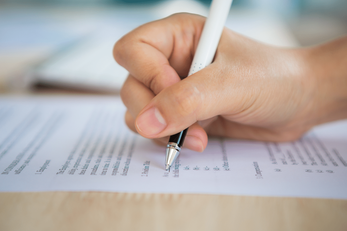 Concursos Públicos  Mão segurando caneta preta preenchendo formulário ou contrato em papel sobre mesa