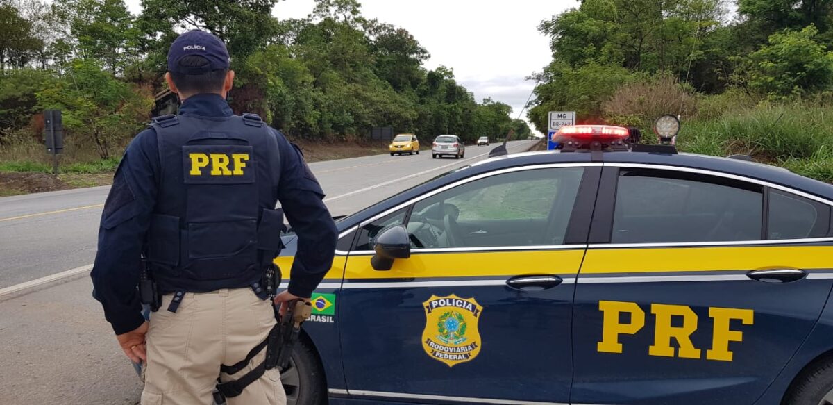 Policial rodoviário federal de costas ao lado de viatura da PRF com luzes piscando na estrada