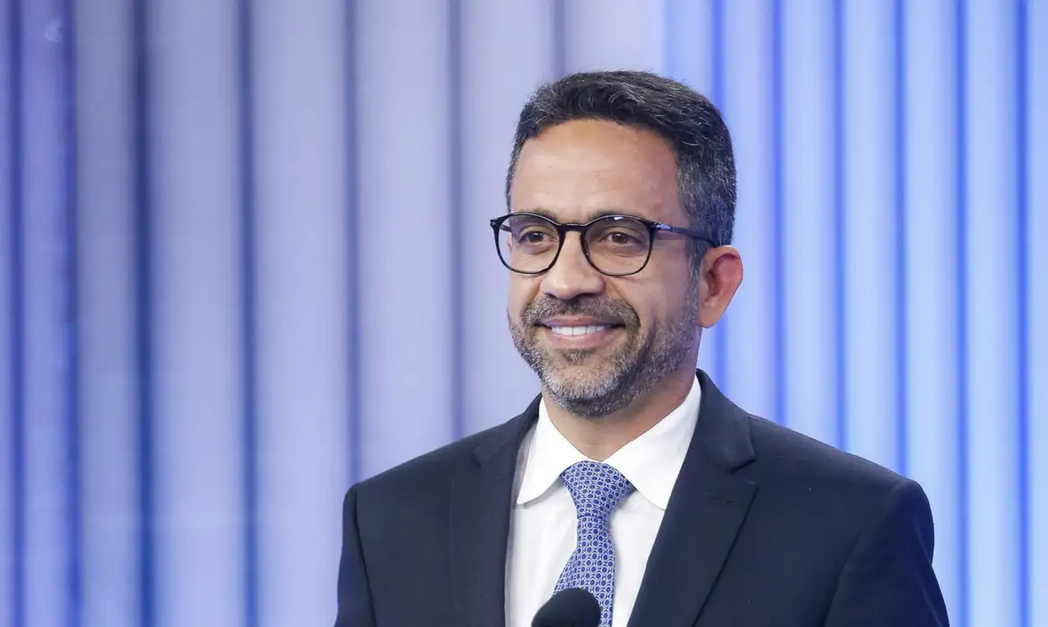 Homem com óculos, terno escuro e gravata azul sorrindo sob luzes azuis desfocadas