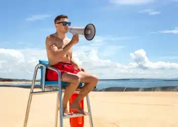 Guarda-vidas com megafone na praia, em posição de vigilância, destacando a oportunidade de trabalho para a Prefeitura com inscrições se encerrando.
