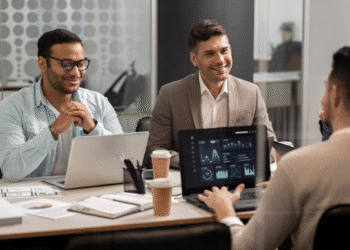 Três homens em reunião de negócios analisando gráficos e dados financeiros em laptops em escritório moderno