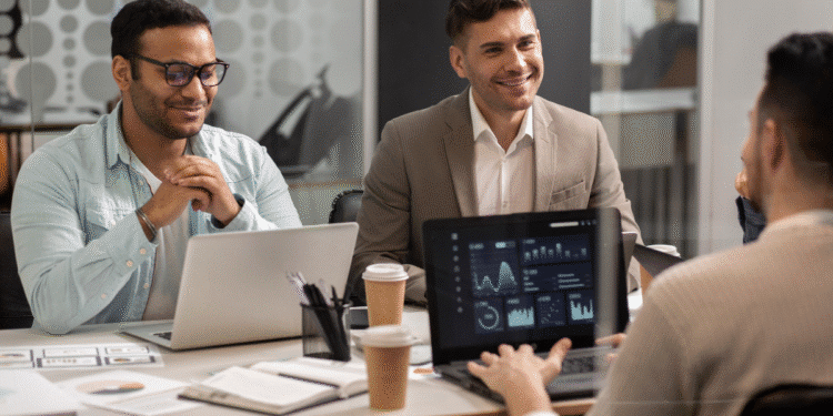 Três homens em reunião de negócios analisando gráficos e dados financeiros em laptops em escritório moderno