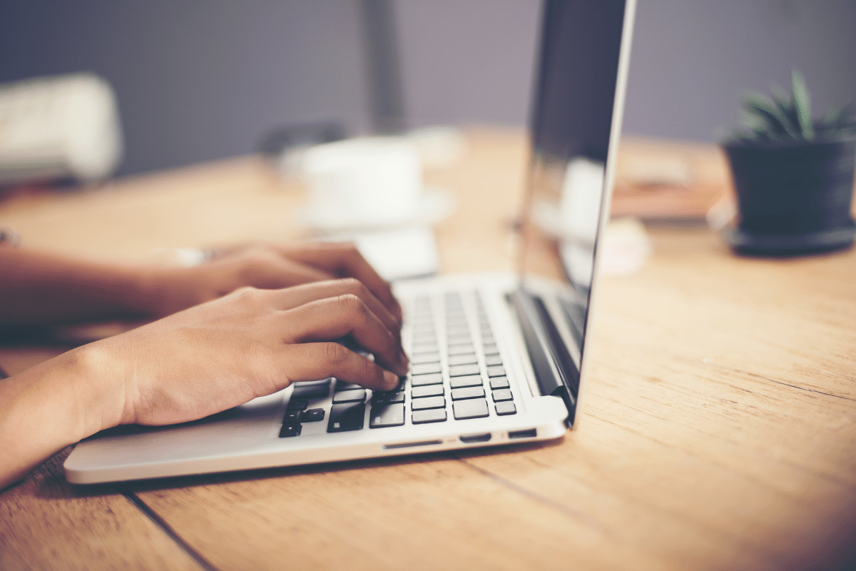 Duas mãos femininas digitando em teclado de laptop prateado sobre mesa de madeira clara.