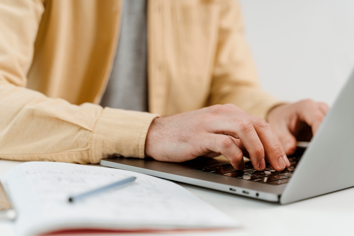 Inscrição concurso Mãos masculinas digitando em teclado de notebook prateado com bloco de notas e caneta ao lado