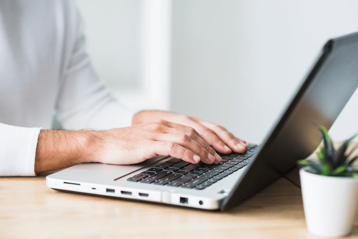 Mãos masculinas digitando em teclado de notebook prateado sobre mesa de madeira clara com planta ao lado