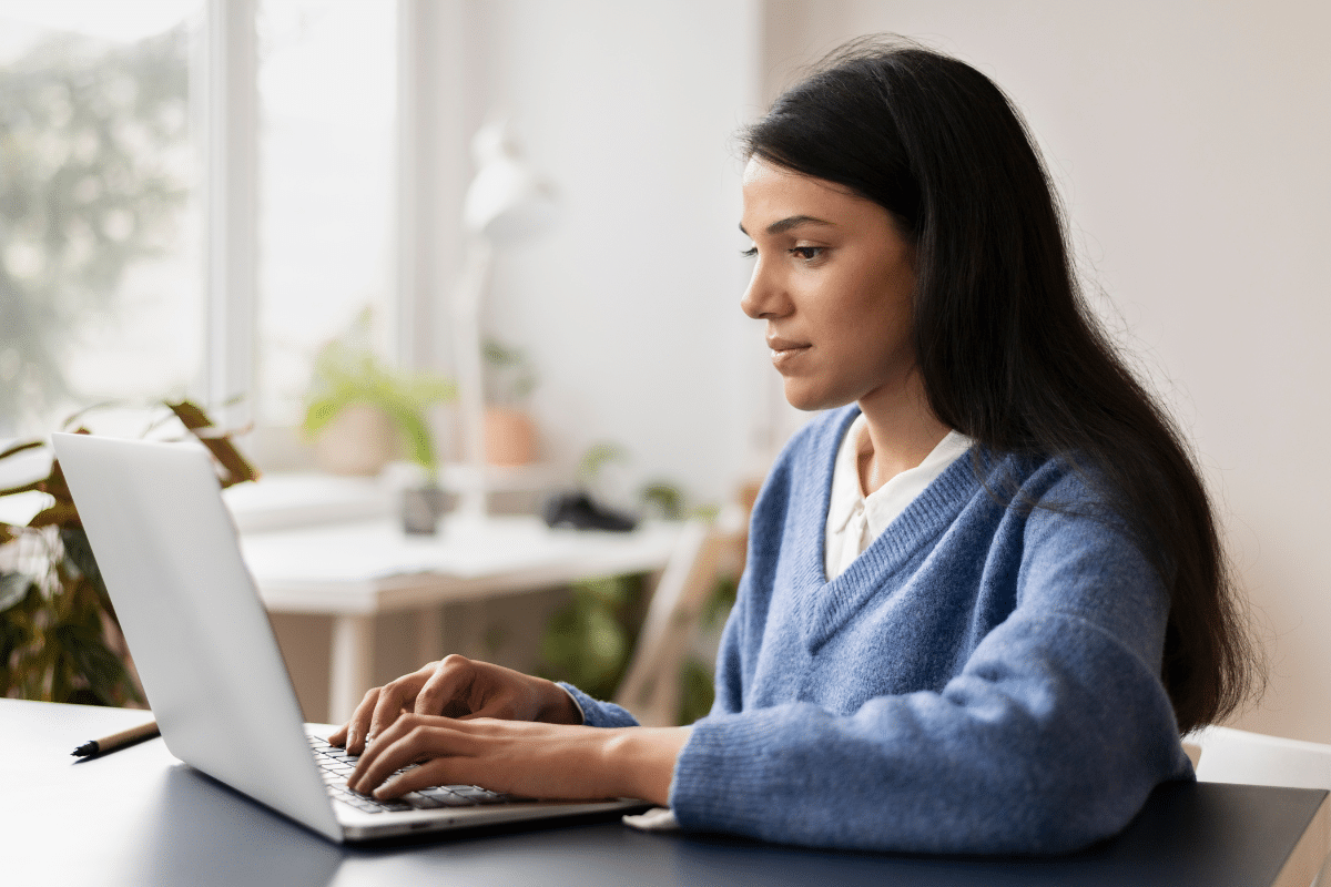Mulher jovem usando suéter azul trabalhando focada em um notebook branco em ambiente claro e moderno