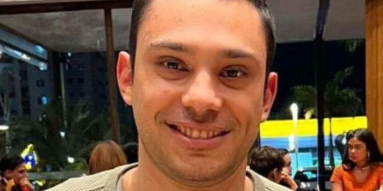 Homem adulto sorrindo, vestindo blusa verde clara, em restaurante com pessoas ao fundo