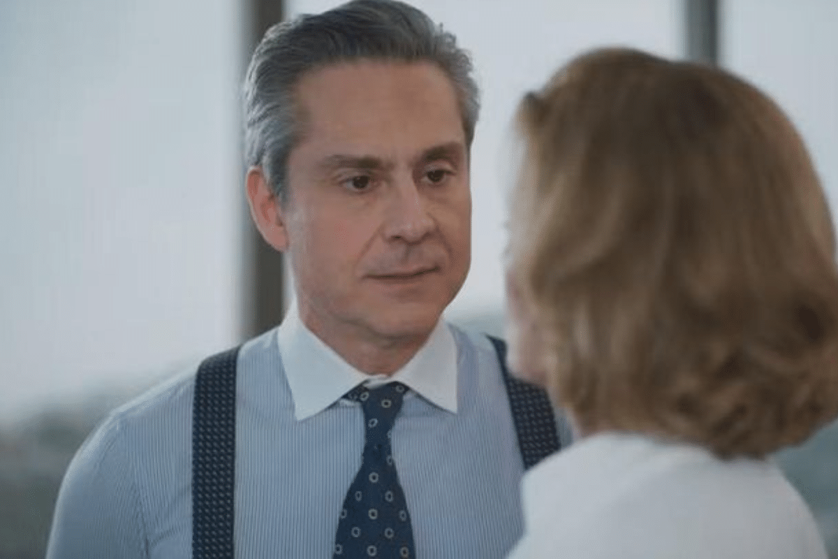 Homem de meia-idade com camisa azul e suspensórios conversando com mulher loira em ambiente de escritório