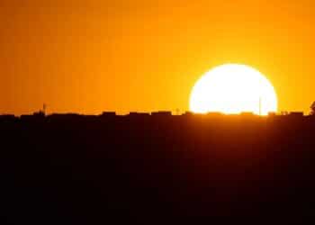 Sol intenso em tons de laranja e amarelo, formando a silhueta de uma cidade, representando a discussão sobre o possível retorno do Horário de Verão em novembro de 2025 e o aproveitamento da luz do dia.