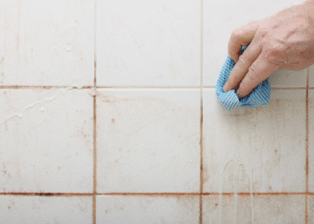 Mão limpando azulejo branco com pano azul listrado para remover sujeira acumulada.