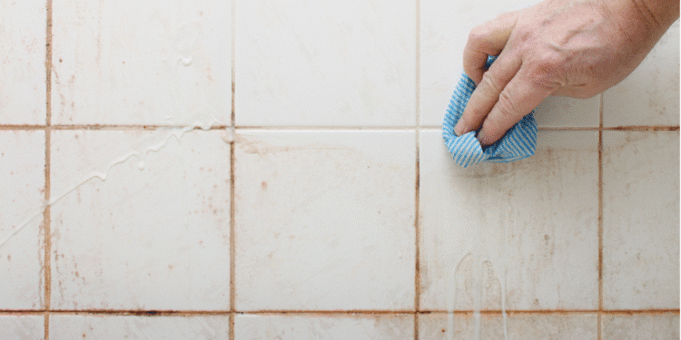 Mão limpando azulejo branco com pano azul listrado para remover sujeira acumulada.