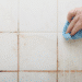 Mão limpando azulejo branco com pano azul listrado para remover sujeira acumulada.