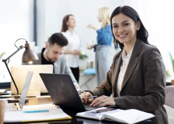 Jovem administradora sorrindo no escritório com laptop, representando as oportunidades de carreira em 2025
