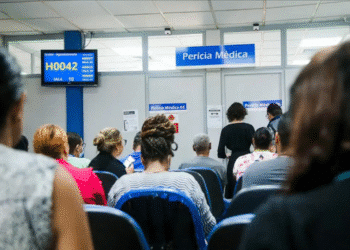 Pessoas aguardando sentadas em cadeiras azuis na sala de espera de perícia médica com painel de senha exibindo H0042