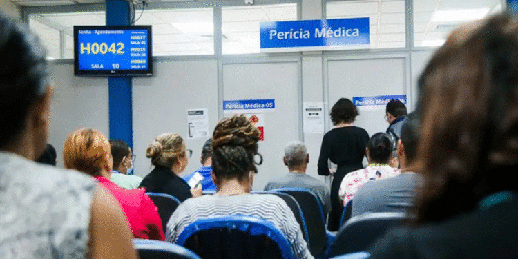 Pessoas aguardando sentadas em cadeiras azuis na sala de espera de perícia médica com painel de senha exibindo H0042