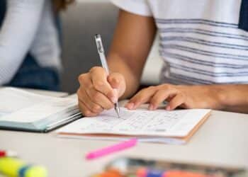 Pessoa preenchendo uma prova, destacando o foco em atividades educacionais, com livros e caneta visíveis, enquanto uma avaliação é realizada.