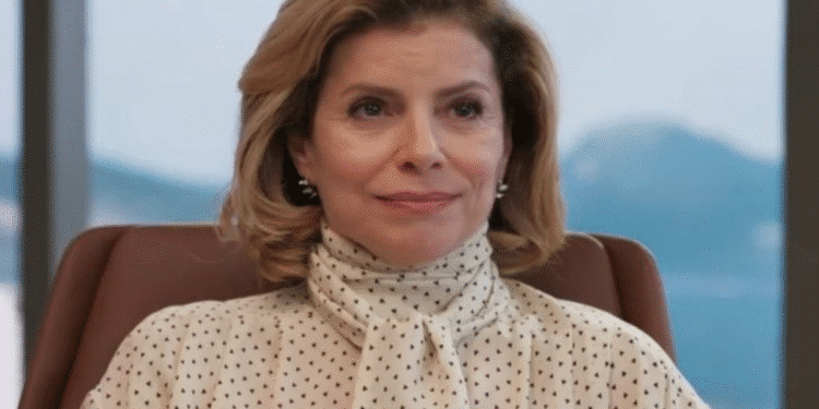 Mulher loira de cabelo curto vestindo blusa branca com poás pretos sentada em cadeira de escritório