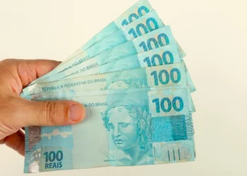 Mão segura cédulas de R$ 100, ilustrando o pagamento de até R$ 1.225 do Pé-de-Meia em agosto.