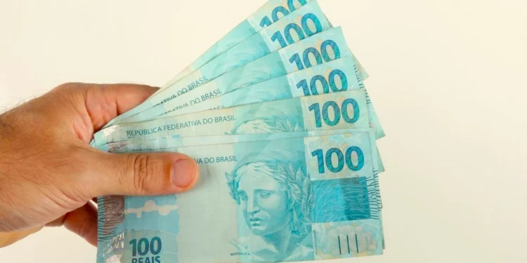 Mão segura cédulas de R$ 100, ilustrando o pagamento de até R$ 1.225 do Pé-de-Meia em agosto.