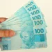 Mão segura cédulas de R$ 100, ilustrando o pagamento de até R$ 1.225 do Pé-de-Meia em agosto.