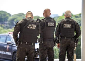 Três agentes da polícia penal vistos de costas com coletes táticos e armas em operação externa