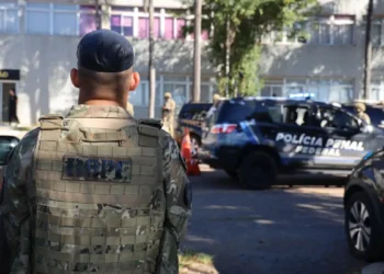 Agente da tropa com uniforme camuflado de costas próximo a viaturas da Polícia Penal Federal em operação